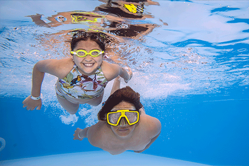 Niño con gafas amarillas y niña con traje de baño de flores y gafas amarillas sonriendo, sumergidos y rodeados de agua, aguantando la respiración aprovechando las promocione de vacaciones Lagomar fin de semana