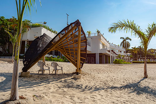 Parasol de madera con techo de lona negro y dos sillas de plástico debajo, ubicado en la arena blanca de la playa de uno de los planes en Lagomar con descuento. Alrededor hay palmeras y en el fondo se ve una construcción blanca con grandes ventanales