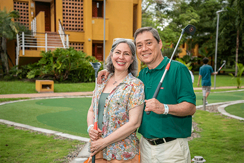Hombre apoyando su mano derecha en el hombro de su pareja mientras disfrutan de la cancha de golf y de los descuentos en hoteles Compensar por compras anticipadas. El hombre vestido con camiseta polo verde y pantalón blanco, y la mujer con blusa verde claro y camisa de flores, sonriendo. En el fondo se ve un edificio de dos pisos color naranja y rejas blancas en las escaleras