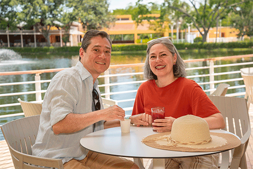 Pareja sonriendo y disfrutando los beneficios de la promoción de planes para ir a Lagomar. Están sentados en una mesa de madera pintada de blanco, con sillas del mismo color, tomando un café y un jugo de mora; el hombre vestido con camisa blanca y la mujer con camiseta naranja y un sombrero sobre la mesa. En el fondo se ve un lago y un edificio rodeado de vegetación