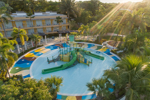 Gran parque para niños de color verde claro y oscuro, ubicado en el medio de la piscina del hotel de dos pisos, con ventanales y paredes de color mostaza en donde puedes encontrar gran cantidad de planes en Lagomar con descuento. Alrededor se ven palmeras y muchos árboles