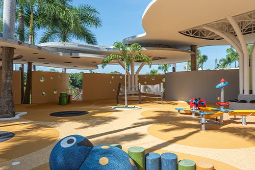 Dos juegos en un parque, el primero de colores amarillo, rojo y verde, y el otro con forma de muñeco azul sonriente acostado al lado de cinco troncos, todo rodeado por un piso amarillo y beige que tiene formas circulares. Alrededor hay techos con formas orgánicas y vista campestre que puedes disfrutar con los descuentos en Lagosol y Lagomar