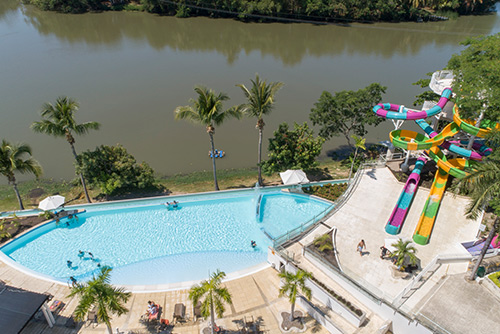Vista panorámica de piscina con agua cristalina en la que hay trece personas dentro disfrutando de la vista al lago y de las promociones de vacaciones Lagomar fin de semana. Alrededor hay un parque con dos grandes toboganes, uno fucsia con azul y el otro amarillo con verde, todo esto acompañado por palmeras, vegetación y parasoles
