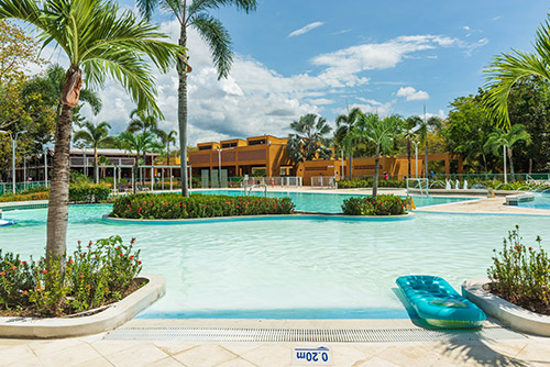 Gran piscina, con pequeñas islas y vegetación, la cual tiene un flotador azul en el borde durante un día soleado. Al fondo de la imagen se ve el hotel de color mostaza, en donde puedes acceder a la promoción reservas anticipadas de Compensar