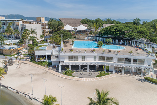 Vista panorámica de la playa con arena blanca y del hotel de dos pisos color blanco que tiene ventanales y una piscina en la que hay varias personas disfrutando del día soleado y de la promoción de reservas anticipadas Compensar. Alrededor hay otros edificios, el mar, palmeras y mucha vegetación