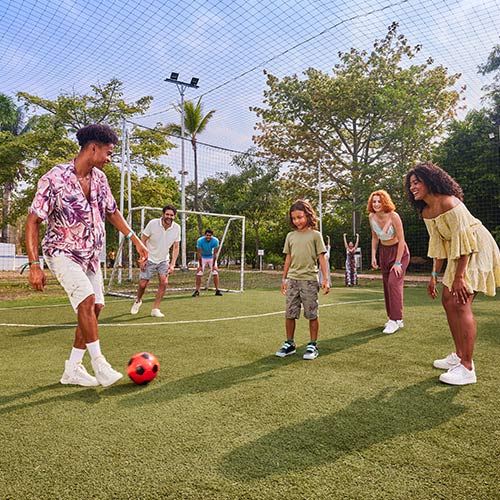 Tres hombres, dos mujeres y un niño, vestidos con ropa para clima cálido de colores blanco, amarillo, verde musgo, morado, vinotinto y azul, están jugando con un balón naranja neón y negro en una cancha de fútbol de Lagomar