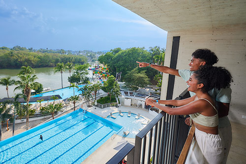 Un hombre y una mujer, que visten ropa para clima cálido de colores azul claro y blanco, están de pie en uno de los balcones del Hotel Lagomar apoyando sus brazos sobre un barandal negro y observando la vista de las piscinas, toboganes y el lago