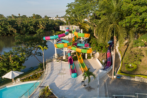Toma aérea de lugar con toboganes incluidos con el plan living viajero Compensar, los cuales son de color morado, azul, amarillo y verde. Alrededor se ve una piscina, un lago y grandes árboles que le dan un toque campestre