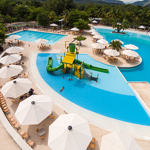Toma aérea de hotel con tres piscinas, en la que se puede ver en el medio un parque con toboganes verdes y amarillos, rodeado con mesas de madera, que tienen parasoles blancos a los cuales puedes acceder con las  promociones de vacaciones en Lagomar fin de semana