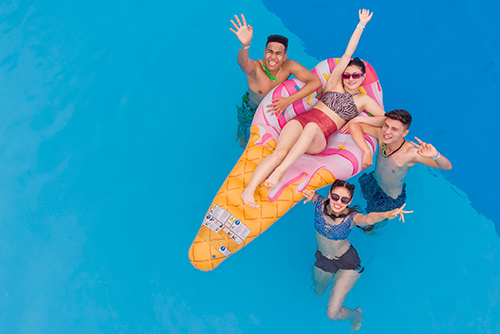 Toma aérea de una piscina en la cual se ven cuatro amigos, dos hombres y dos mujeres, una de ellas sobre un flotador con forma de helado, mientras comparten en el agua y disfrutan uno de los beneficios del plan living viajero Compensar