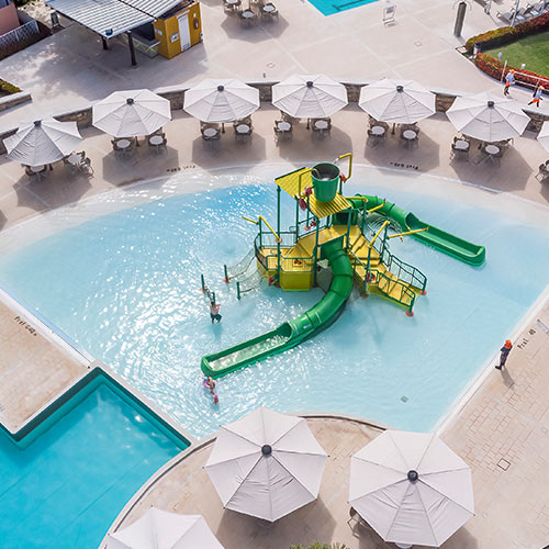 Piscina en temporada baja Lagosol, en su centro hay un parque acuático infantil color verde y amarillo, donde tres personas se divierten y a su alrededor hay mesas con sombrillas de color blanco