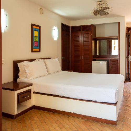 Habitación con cama doble de sabanas y almohadas blancas, mesa de noche y closets de madera que están incluidos en el paquete turístico para viajar a Girardot en semana santa. Las paredes son blancas y tienen dos bombillos que ayudan a iluminar y a resaltar la decoración de un cuadro de colores que hay en la pared, en el techo un pequeño ventilador
