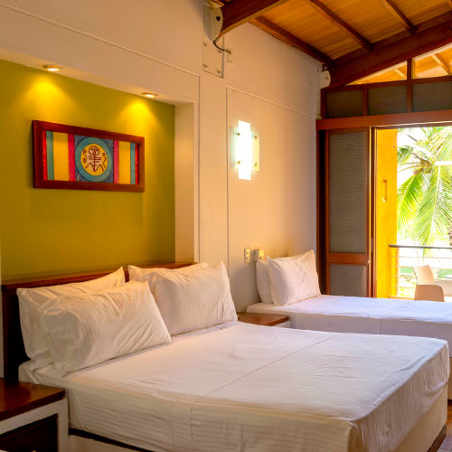 Habitación con dos camas de sabanas blancas y almohadas, con dos mesas de noche. La pared es verde limón y tiene un cuadro de figuras abstractas amarillas, rojas, azules, verdes con marco café. Esta es una de las habitaciones con vista exterior que se encuentran en los planes de semana santa Lagosol y Lagomar. Afuera se ve una palmera y sillas en medio de un día soleado