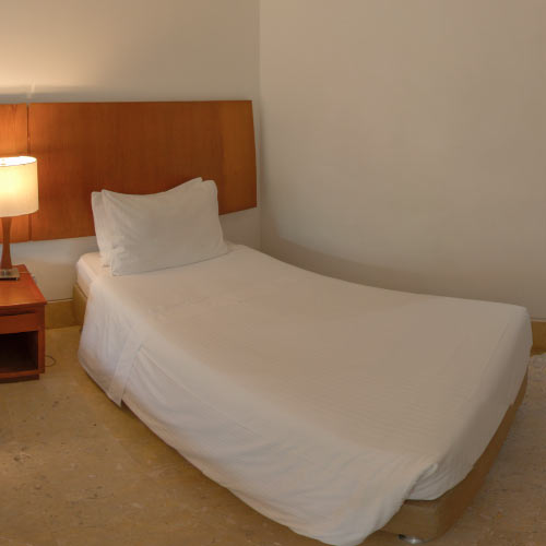 Habitación de cama sencilla con sabanas blancas, espaldar acolchonado anaranjado, mesa de noche y una lámpara encendida, que se pueden encontrar en la oferta de hoteles en Girardot para viajar en semana santa