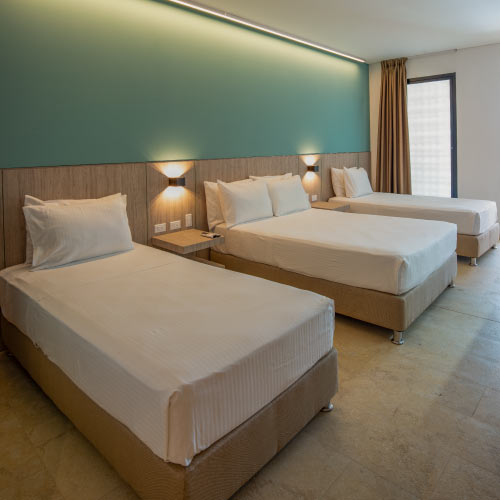 Habitación con dos camas sencillas, una doble, dos mesas de noche, dos lámparas, con detalles de madera en el espaldar, una pared verde limón, una blanca y una gran ventana acompañada de una cortina