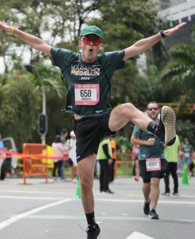 Atleta, que usa camiseta, gorra verde, gafas deportivas rojas y pantaloneta negra, se encuentra en medio de una calle, durante la Maratón en Medellín, con los brazos extendidos y una pierna levantada, como signo de celebración de vivir esta experiencia