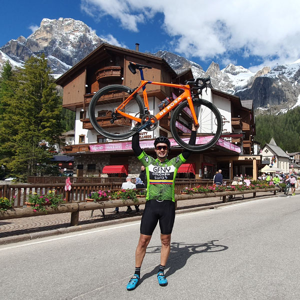 Ciclista levantando su bicicleta en signo de celebración por haber llegado a la meta en medio de uno de los tours para viajar a Europa. Alrededor se ve una casa de tres pisos, árboles y una gran montaña con nieve que se mezcla con las nubes
