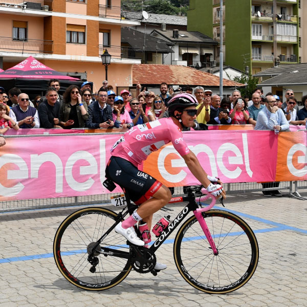 Hombre que usa camiseta rosada, short negro, monta su bicicleta de camino a la meta de una de las competencias de ciclismo incluidas en el paquete de viaje para el tour de Francia. Alrededor se ve una gran cantidad de personas observando y disfrutando del evento