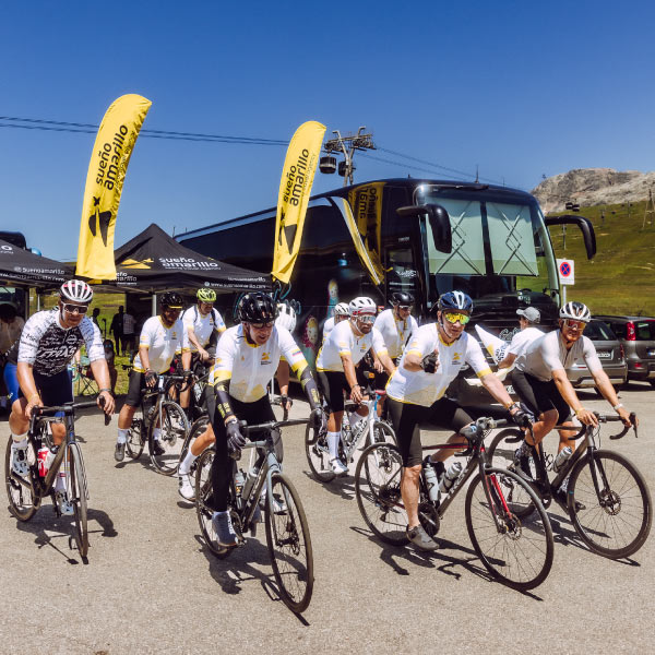 Grupo de ciclistas, vestidos con uniforme blanco con negro, iniciando la competencia que hace parte del paquete de viaje giro de Italia. Alrededor se ve un bus y carpas negras, acompañados de dos banderas amarillas que tienen escrito en negro “sueño amarillo”