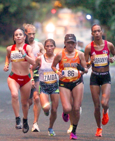 Cuatro corredoras y un corredor viven una de las mejores carreras atléticas para runners, mientras avanzan por una calle rodeada de arboles y luces de carros en el fondo