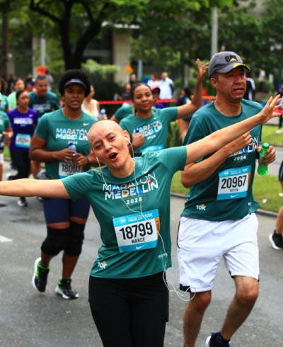 Corredora rapada con los brazos extendidos, usa una camiseta verde en la que se ve el número 18799 que corresponde a su identificación como participante de la Maratón en Medellín. Detrás de ella otros corredores buscando llegar a la meta