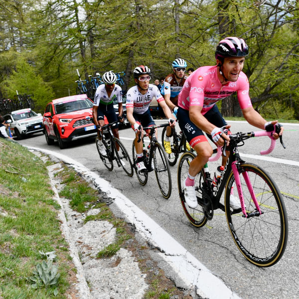 Cuatro personas pedaleando por una carretera empinada son escoltados por un vehículo rojo, uno negro y uno gris, los cuales hacen parte del paquete de viaje para el tour de Francia. Alrededor hay gran cantidad de naturaleza y algunos espectadores que disfrutan de la competencia