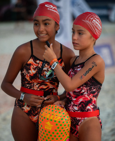 Dos niñas, en medio de la playa, que usan traje de baño. una de color negro, naranja y blanco y la otra negro, rojo y blanco y en su cabeza un gorro de color rojo Turismo deportivo Compensar Plan plus swimmer