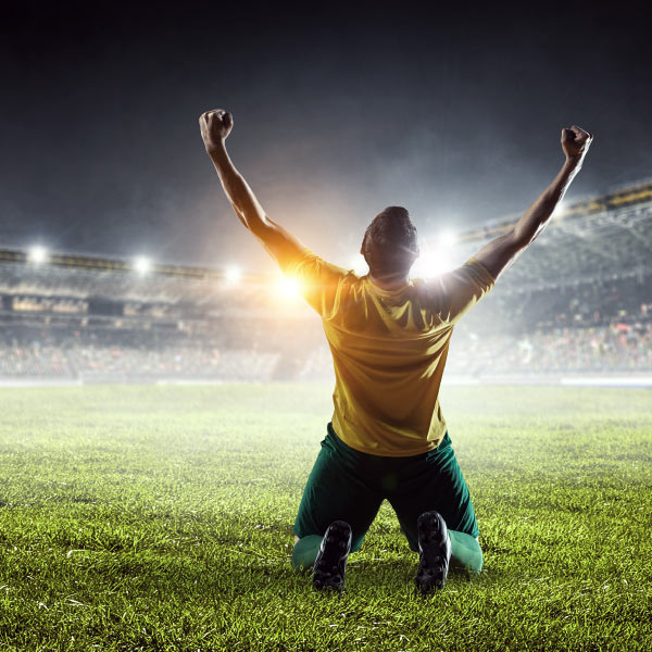 Hombre arrodillado en el pasto de la cancha de un estadio de fútbol, con los brazos levantados en modo de celebración, durante un partido que se puede disfrutar con la oferta de turismo deportivo Compensar