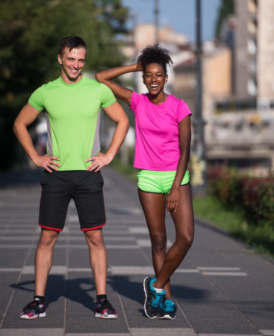 Hombre alto, con camiseta verde neón y pantaloneta negra, se encuentra al lado de una mujer morena, que usa camiseta fucsia y short verde neón, la cuál apoya el codo en el hombro de su compañero. Los dos están en medio de la calle listos para la media maratón Cali