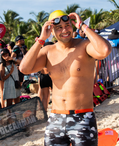 Hombre en medio de la playa que está sonriendo, usa gorro de baño amarillo y gafas de natación en una de las locaciones rodeadas de palmeras incluidas con las ofertas de turismo deportivo. En el fondo se ven varias personas observando en evento de natación