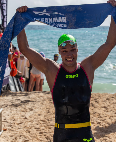 Hombre, con los brazos levantados hacia arriba, sostiene una bandera de color azul durante competencia Oceanman, la cuál se lleva a cabo en la playa y está incluida dentro de las ofertas de turismo deportivo de Compensar. En el fondo se ve un grupo de personas reunidas cerca al mar