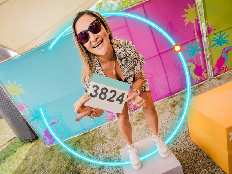 Mujer rubia con lentes de sol, sonriendo sobre un cubo gris, muestra su numero de competencia que obtuvo al adquirir planes en oferta para correr maratones en Colombia con Compensar. En fondo se ve una pared de colores fucsia, cian y verde