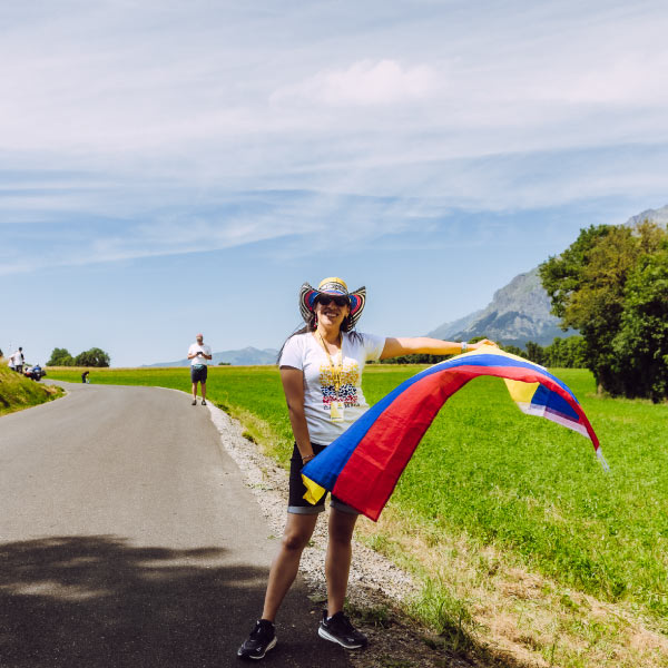 Mujer con camiseta blanca, short negro y un sombrero típico, sostiene una bandera de Colombia en medio de una carretera que está rodeada de árboles y naturaleza, mientras vive y disfruta de los beneficios de comprar boletos para eventos deportivos de Compensar