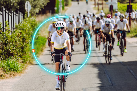 Mujer vestida con uniforme blanco y short negro montando bicicleta durante una competencia luego de acceder a uno de los paquetes de turismo deportivo en Compensar. Detrás de ella hay gran cantidad de ciclistas que usan el mismo uniforme, mientras pedalean por una vía rodeada de naturalezao