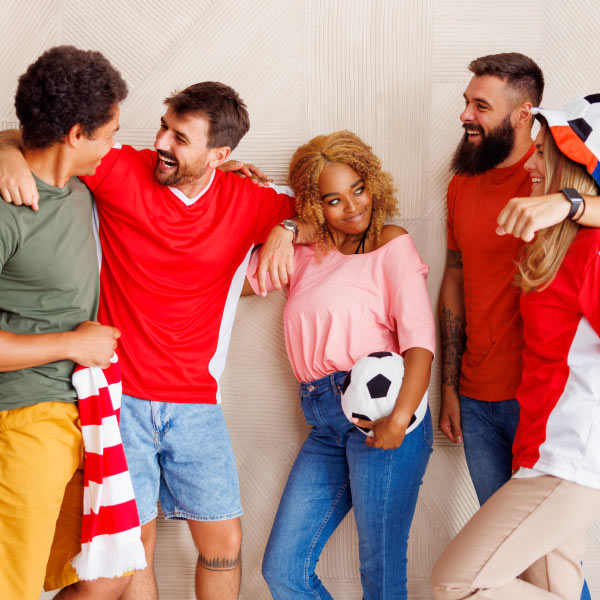 Tres hombres y dos mujeres están compartiendo y sonriendo al descubrir todas las ofertas de turismo deportivo disponibles en Compensar. Las personas usan ropa casual, una de ellas sostiene un balón y su compañero sostiene una bufanda de líneas de color rojo y blanco