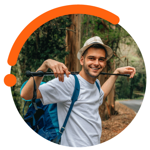 Un hombre, que utiliza un sombrero de cuadros, camiseta blanca y mochila azul, está de pie sosteniendo un palo de trekking de color negro sobre sus hombros y apoyando sus manos en cada extremo, a su alrededor hay diferentes tipos de árboles y un sendero