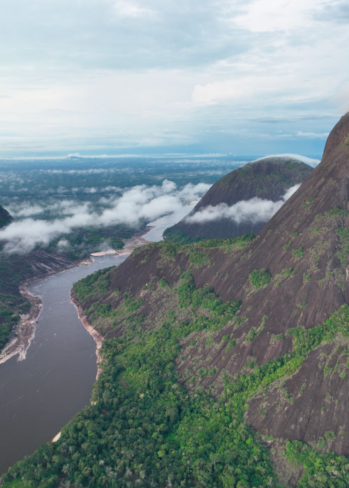 Toma aérea de un río y de los cerros de Mavicure, en los cuales se alcanza a ver vegetación y tierra de color café, los que hacen parte de los mejores lugares para hacer ecoturismo en Colombia