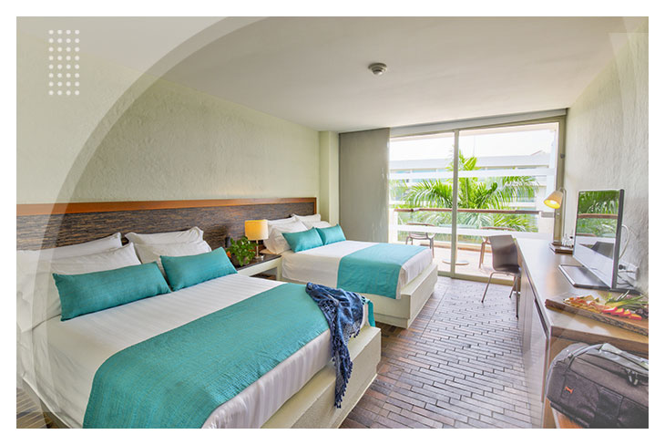 Habitación amplia con dos camas, cobijas color azul, escritorio, televisión, y balcón con vista a las palmeras, perfecta para descansar en el Hotel Estelar Playa Manzanillo Cartagena.