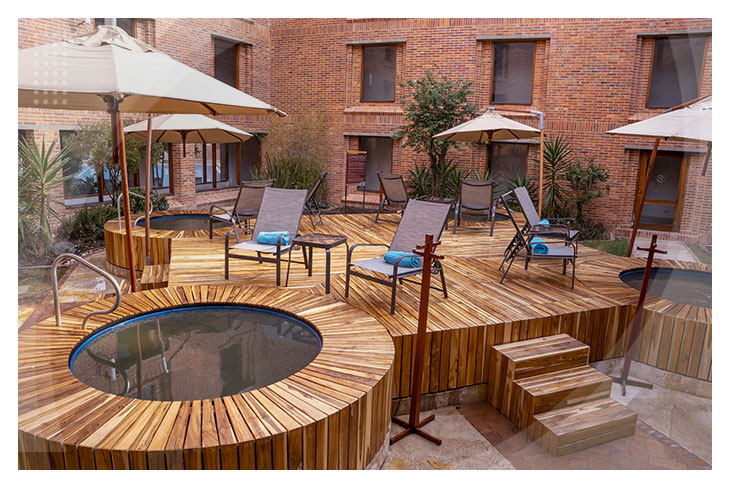 Zona de descanso en terraza de madera con sillas, sombrillas y pequeños jacuzzis al aire libre rodeados de un edificio de ladrillo en un hotel cerca al lago Sochagota.