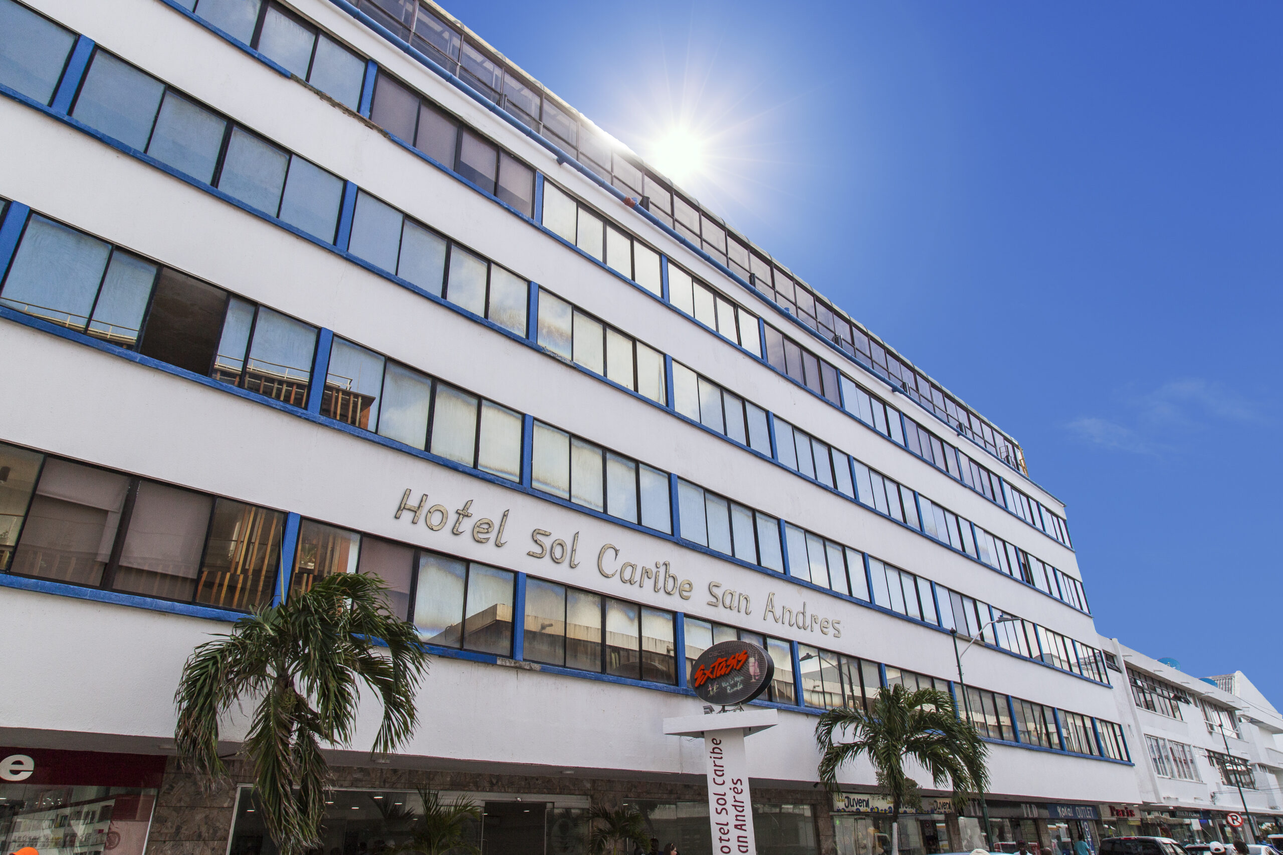 Edificio blanco de varios pisos con ventanales amplios y palmeras al frente, correspondiente al Hotel Sol Caribe San Andrés, ubicado en una zona urbana cercana al comercio y servicios de la isla
