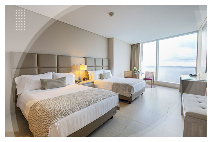 Habitación doble con diseño moderno, camas con sábanas blancas, ventanal de piso a techo con vista al mar y muebles en tonos claros, ideal para quienes buscan hoteles cerca al mar en Cartagena.