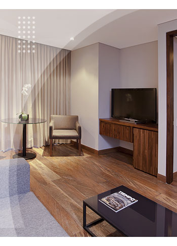 Sala de habitación con piso en madera, sofá gris, mesa de centro, televisor y un rincón acogedor con sillas y cortinas largas es parte de los espacios modernos del Hotel Estelar del Parque de la 93.