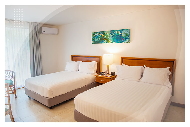Habitación con dos camas, decoración sencilla en tonos claros, mesa de noche con lámpara y acceso a un balcón, en un hotel cerca a la playa Pozos Colorados.