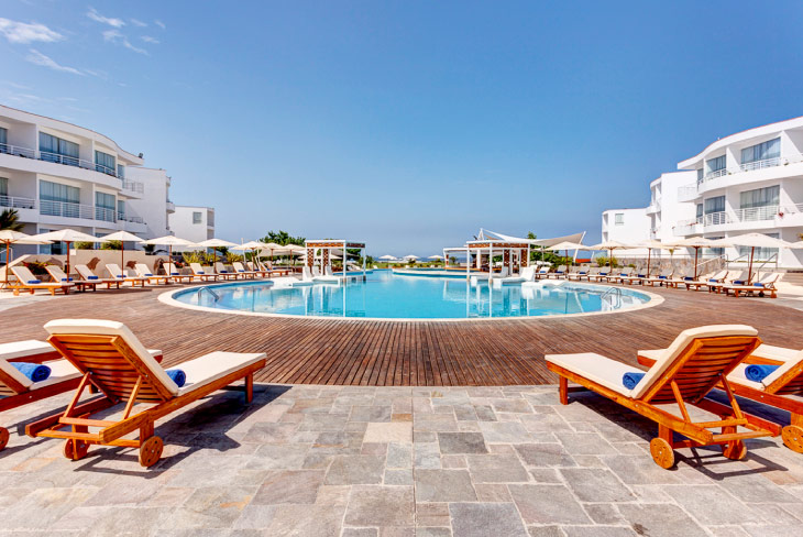 Piscina al aire libre rodeada de sillas asoleadoras de madera y edificios blancos, parte de las ofertas de los hoteles Estelar Perú para disfrutar zonas comunes amplias y luminosas